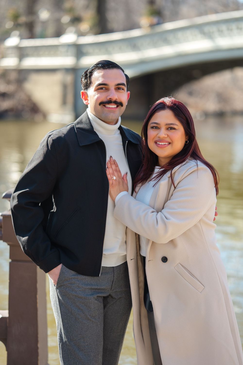 A romantic couple poses together on a bridge wearing coordinated winter coats in cream and black.