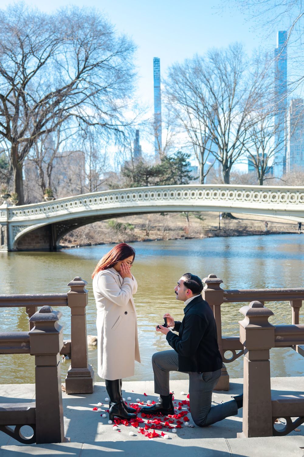A couple gets engaged during winter at Bow Bridge in Central Park with tall buildings visible in the background.
