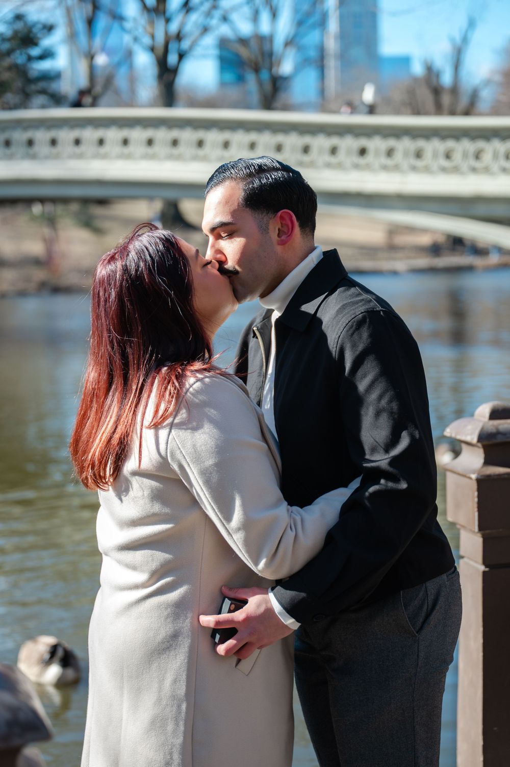 A romantic couple shares a kiss by Bow Bridge in Central Park with a serene lake and winter trees in the background after proposal