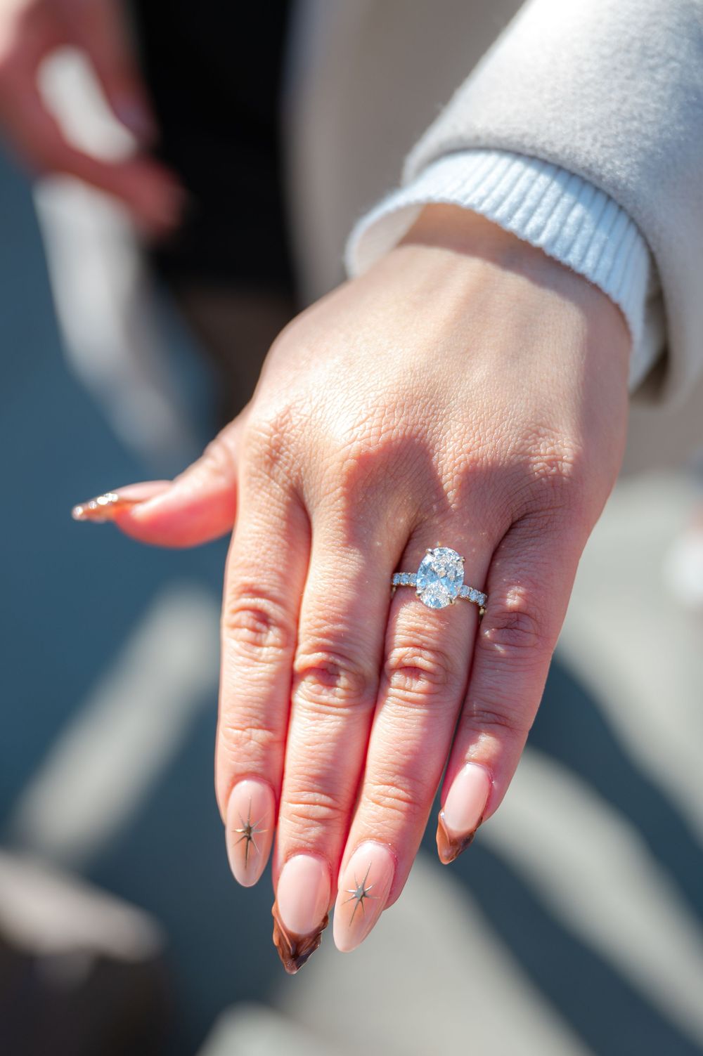 A close-up of a sparkling diamond engagement ring on a manicured hand with pink nail polish.