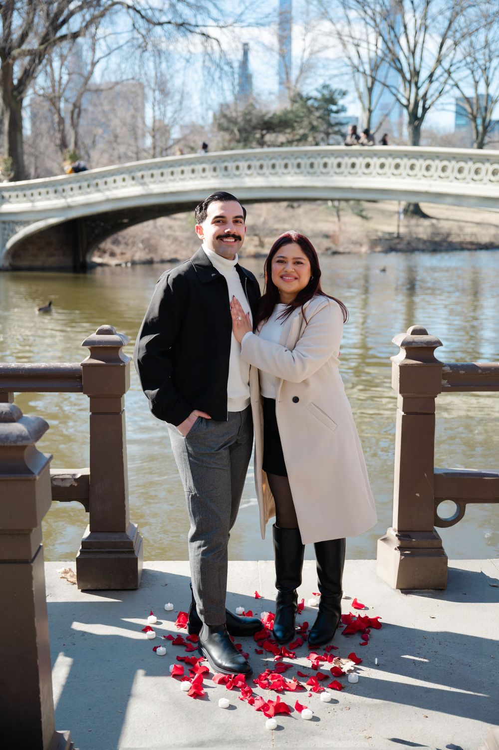 A happy couple poses on a bridge with red rose petals at their feet after surprise proposal in Central Park in NYC