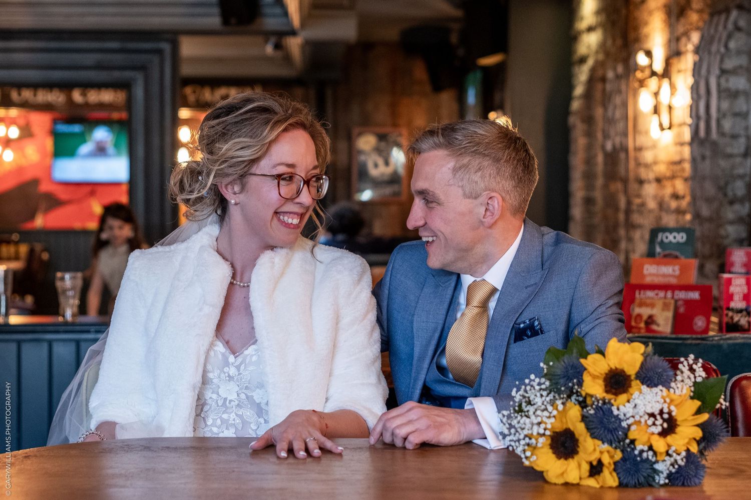 Happy couple shares intimate moment at rustic restaurant table with sunflowers and white fur wrap on wedding day.