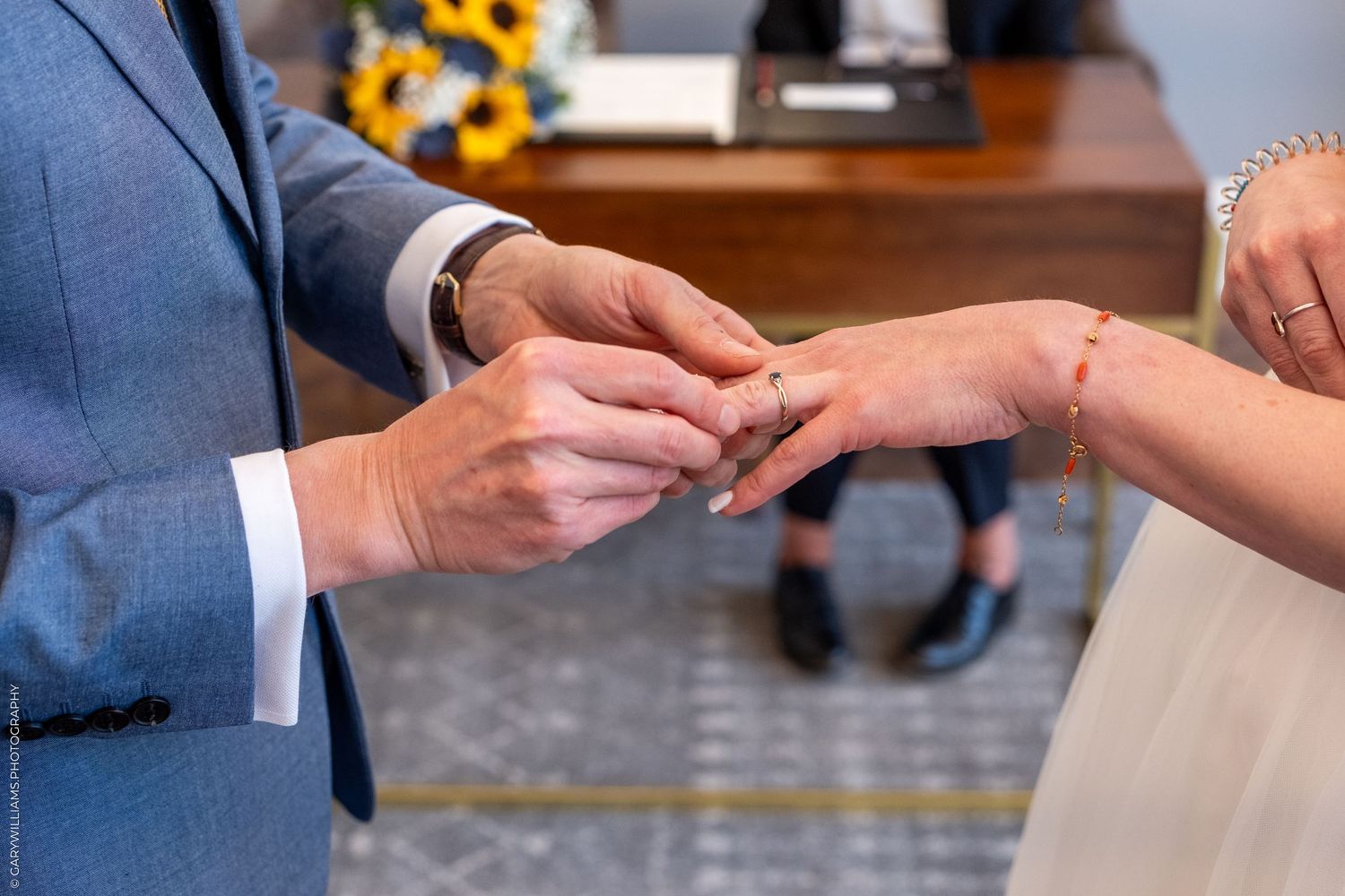 Close-up view of hands exchanging wedding rings during an intimate ceremony with sunflowers in the background.