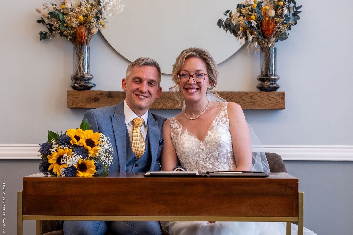 Two people sign documents at a wooden desk while smiling, with yellow sunflower bouquets and floral arrangements decorating the scene.