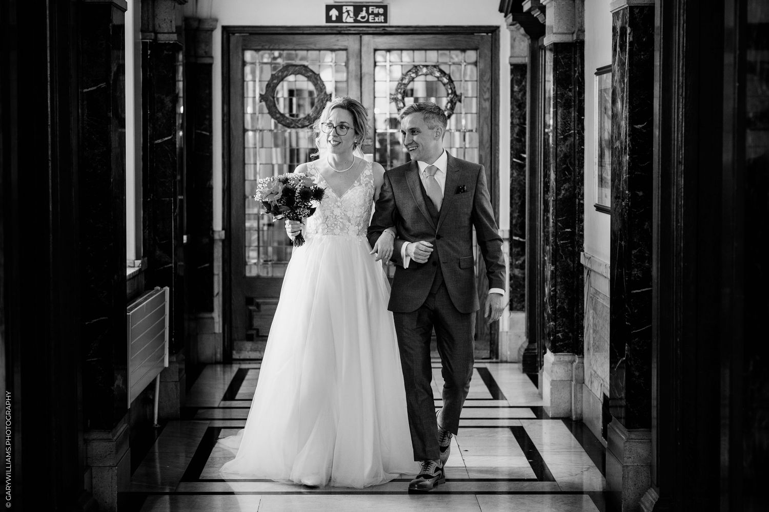 A black and white wedding photo showing a couple walking arm in arm down an elegantly decorated hallway.