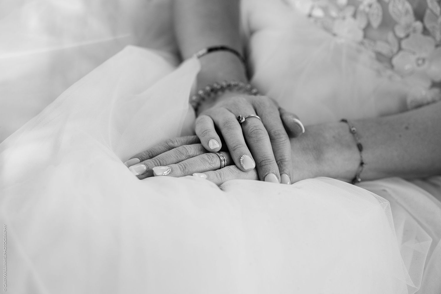 Detail of hands wearing rings resting on white wedding dress fabric in black and white photography.