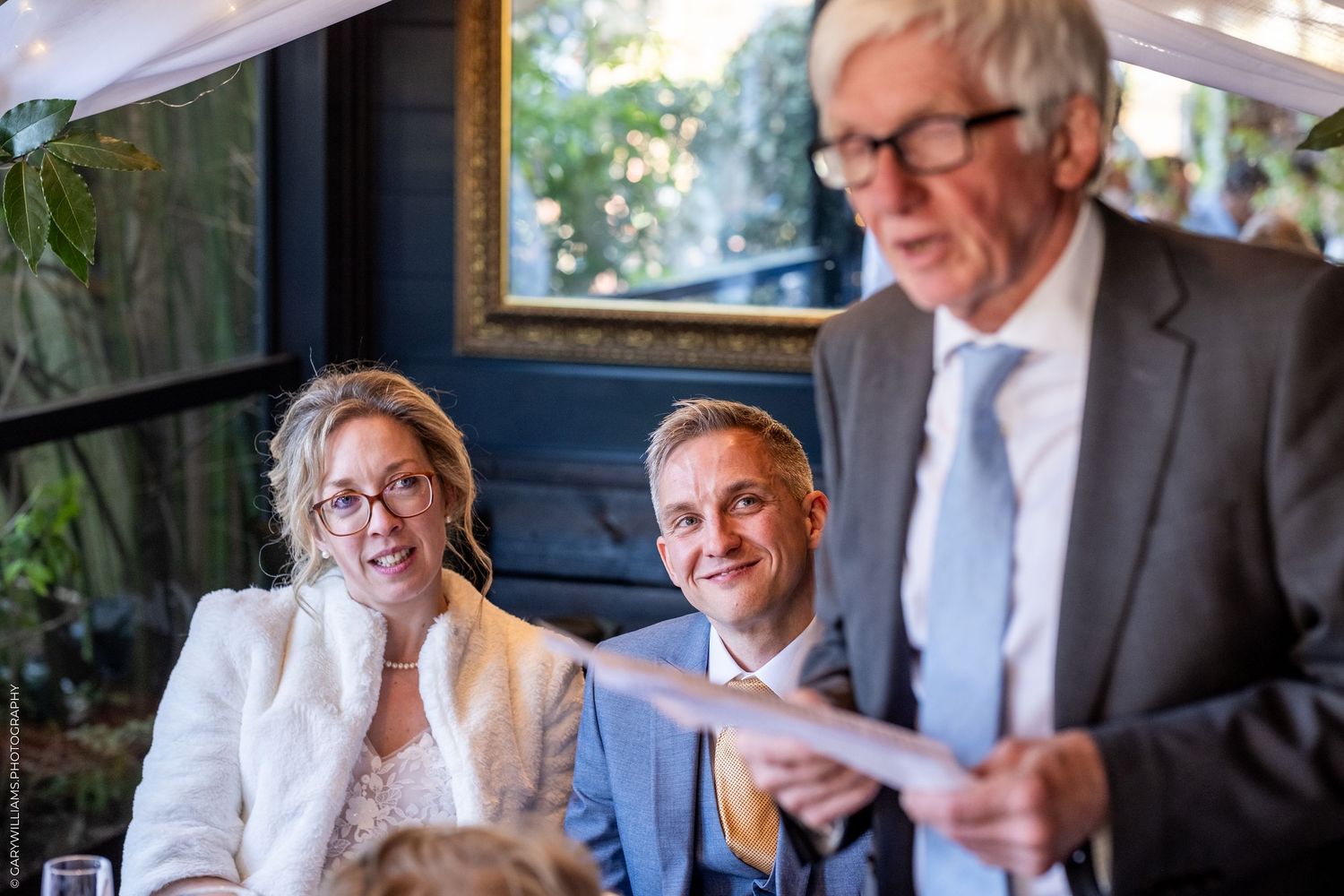 A happy couple sits together at a restaurant table while an older person in a suit reads during a celebration event.