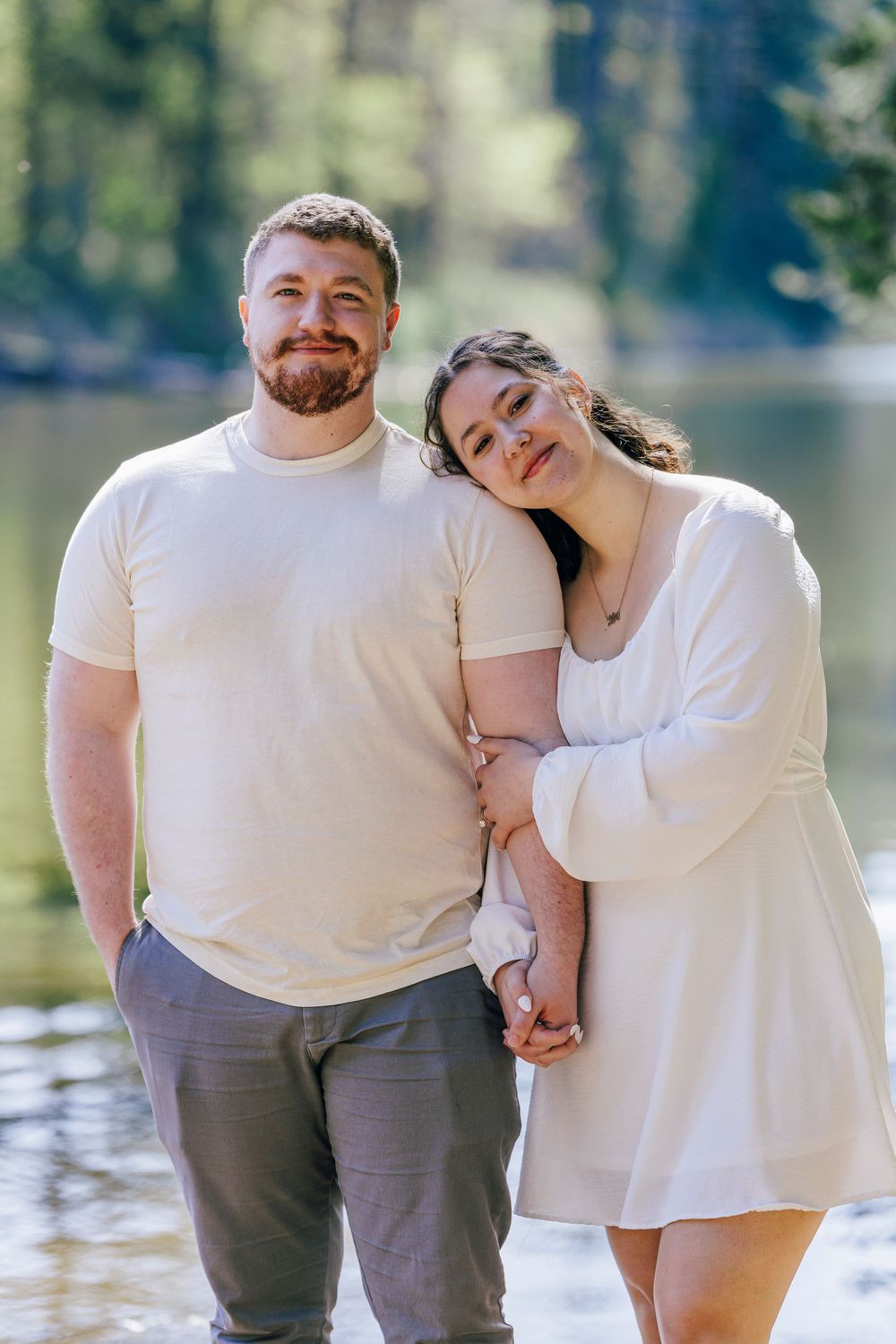 A couple in matching white outfits poses together by a lake on a sunny day.