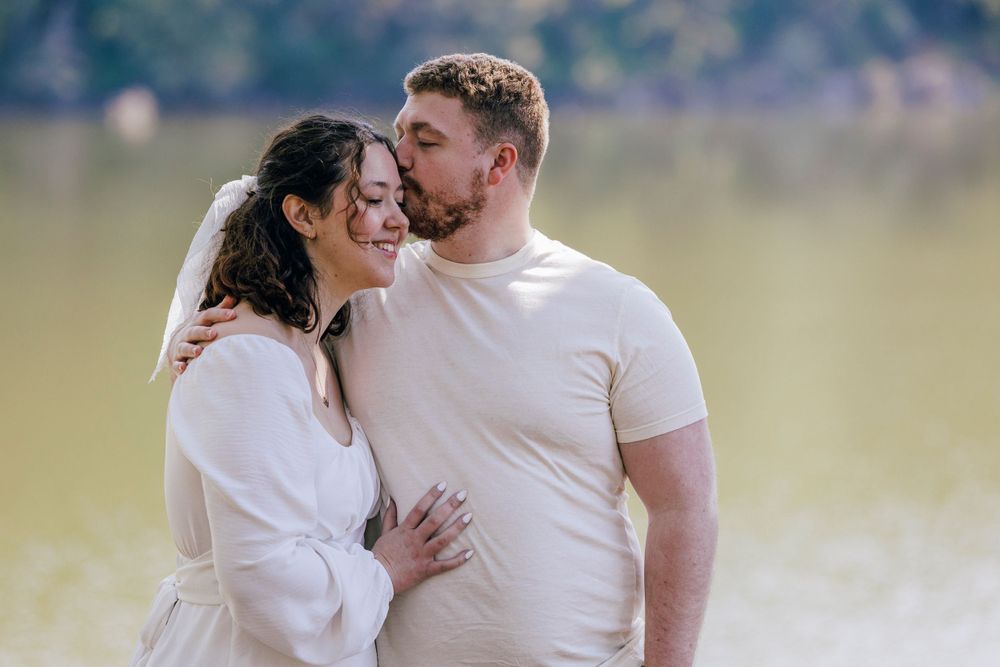 A couple in white clothing embrace lovingly while standing in a sunlit meadow near a lake.