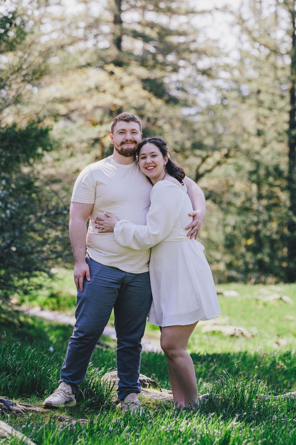 A romantic couple wearing coordinated white outfits embraces while standing in a sunny forest clearing.