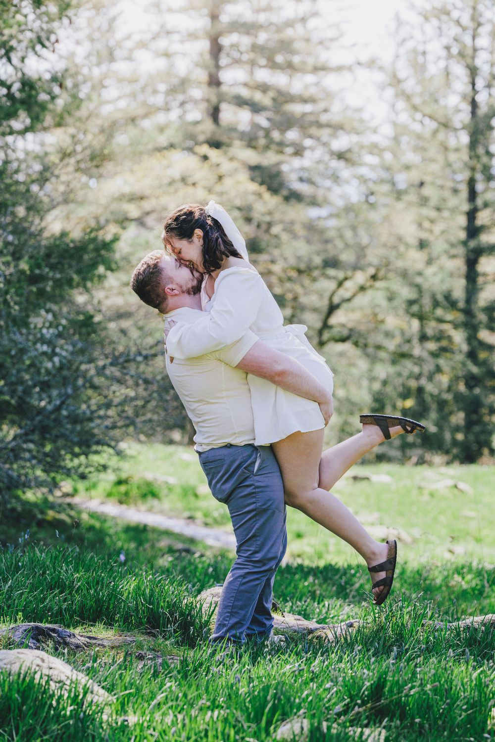A couple in white embraces playfully on a grassy hillside surrounded by evergreen trees.