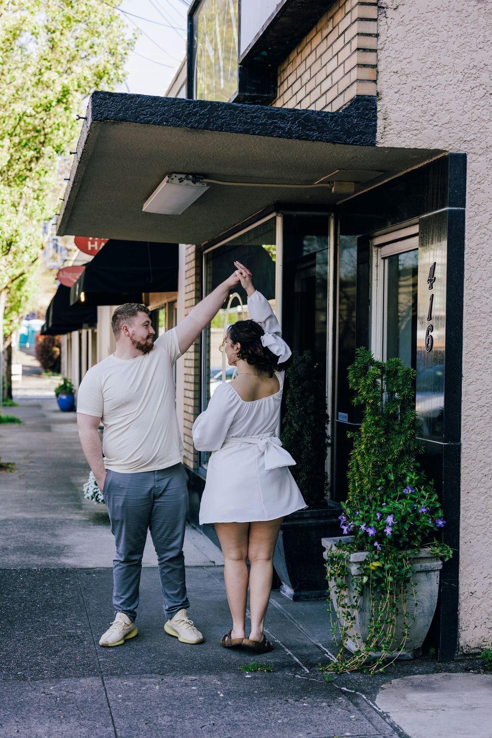 A couple poses playfully outside a brick building with an awning.
