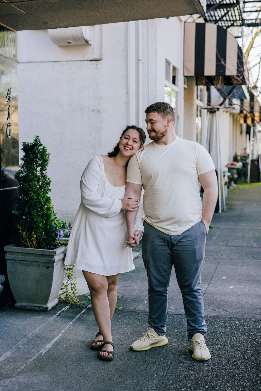A couple walks together along a sidewalk outside a building, wearing casual white outfits on a cloudy day.
