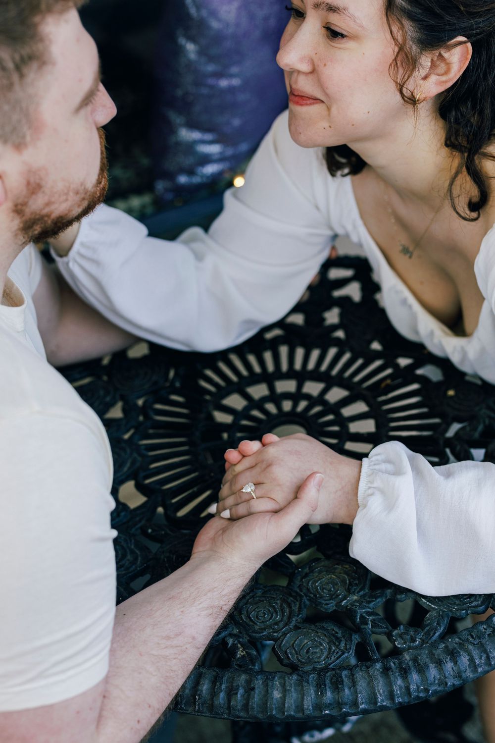 A couple in white clothing holds hands intimately across a decorative black metal table.