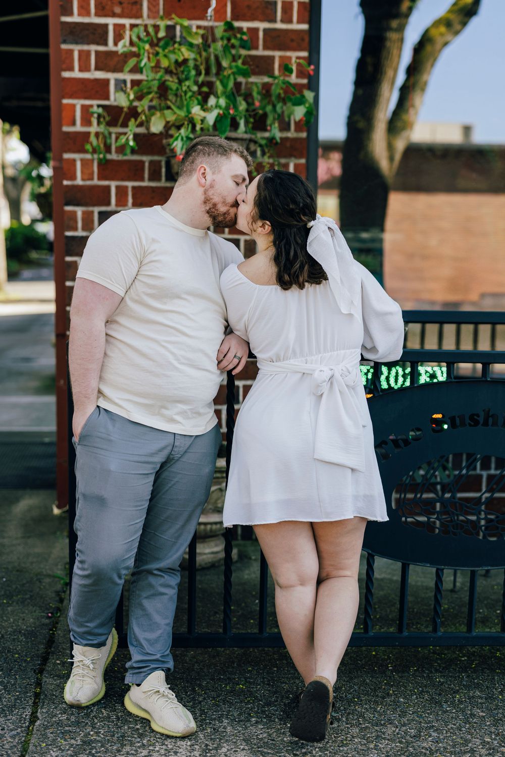 A couple in coordinated white outfits poses together against a brick building backdrop.