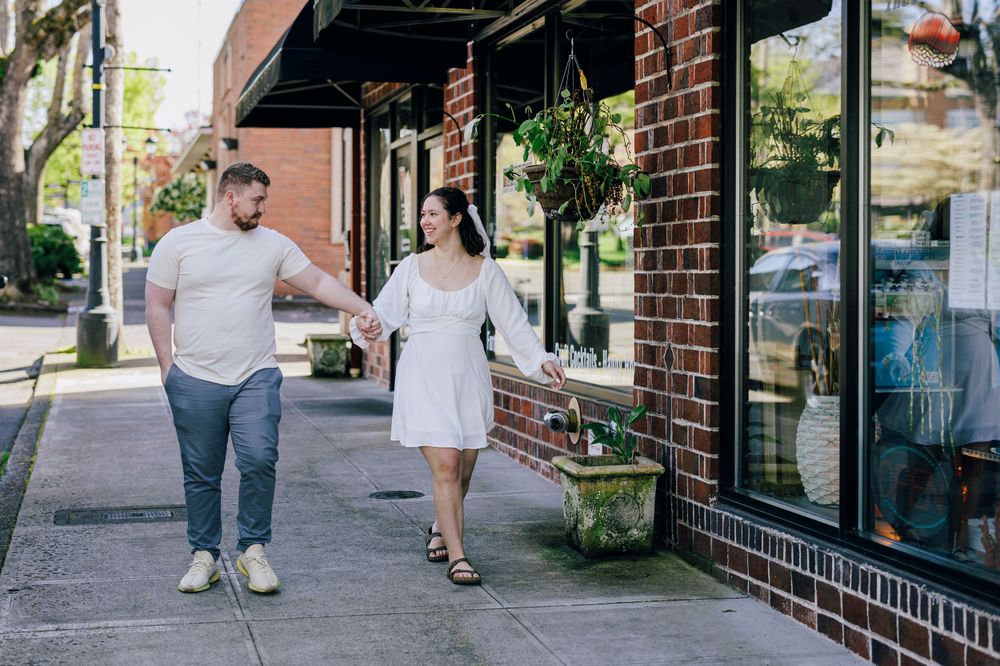 A couple walking together on a sidewalk past brick storefronts in an urban setting.
