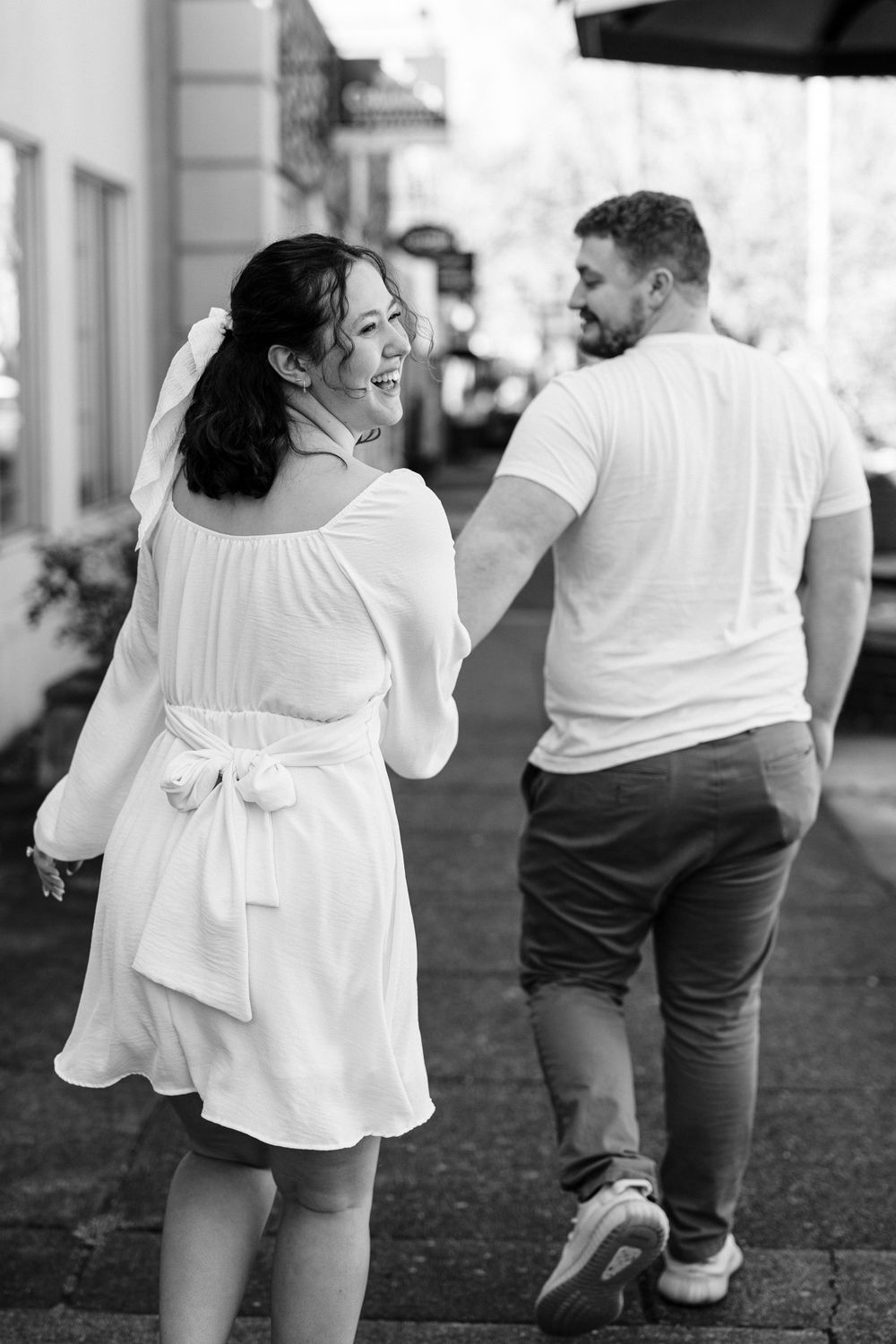 A couple walking together on a street, with her in a flowing white dress and him in casual shorts and shirt.