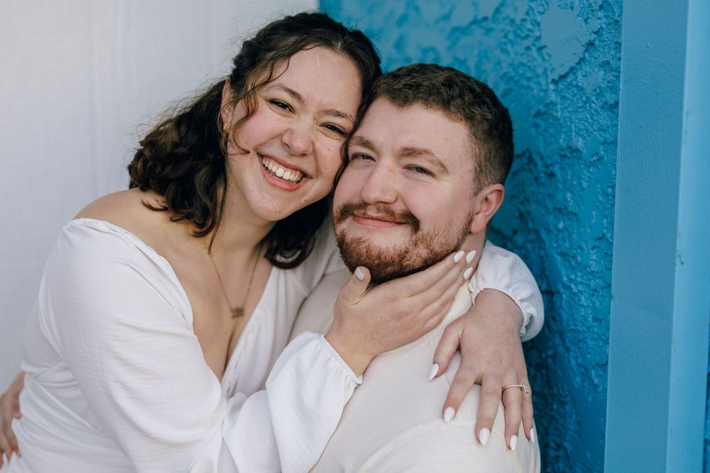 A joyful couple wearing white embraces while posing against a vibrant turquoise wall.