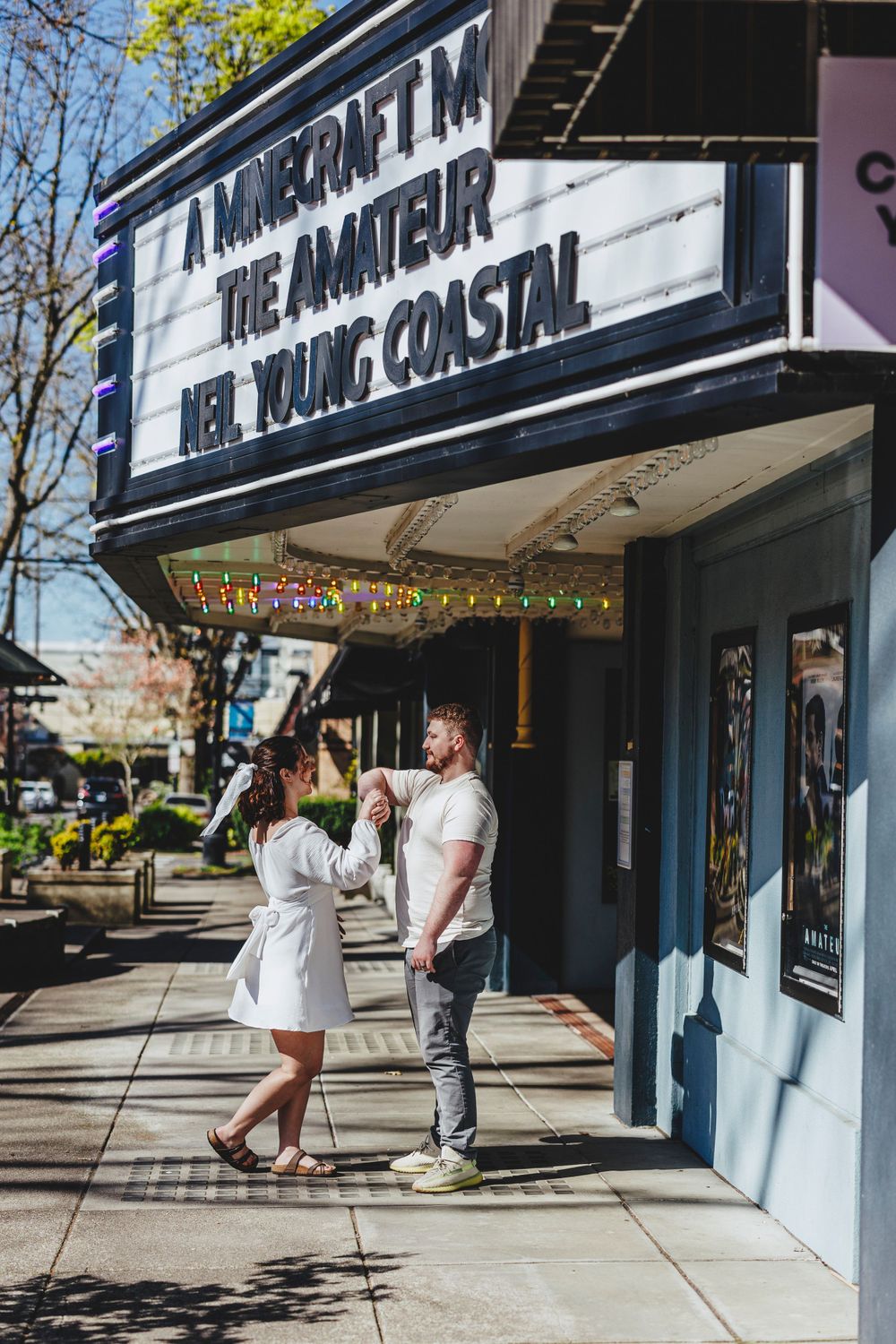 A couple stands outside a movie theater with a marquee reading 'A Murder In The Amateur' on a sunny street.