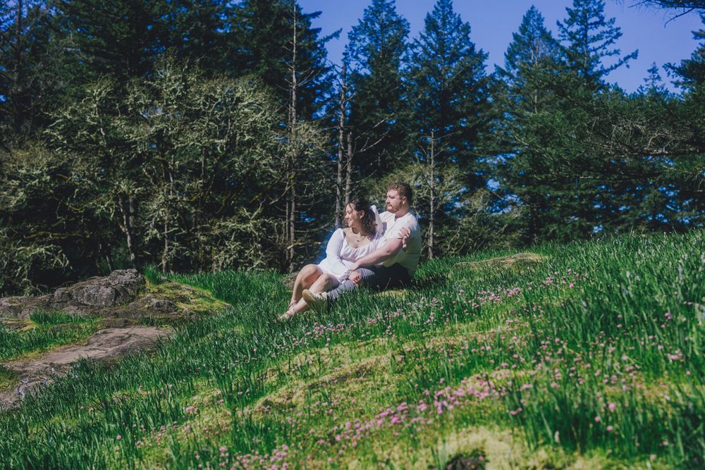 Two people sit together on a grassy hillside path surrounded by evergreen trees on a sunny afternoon.