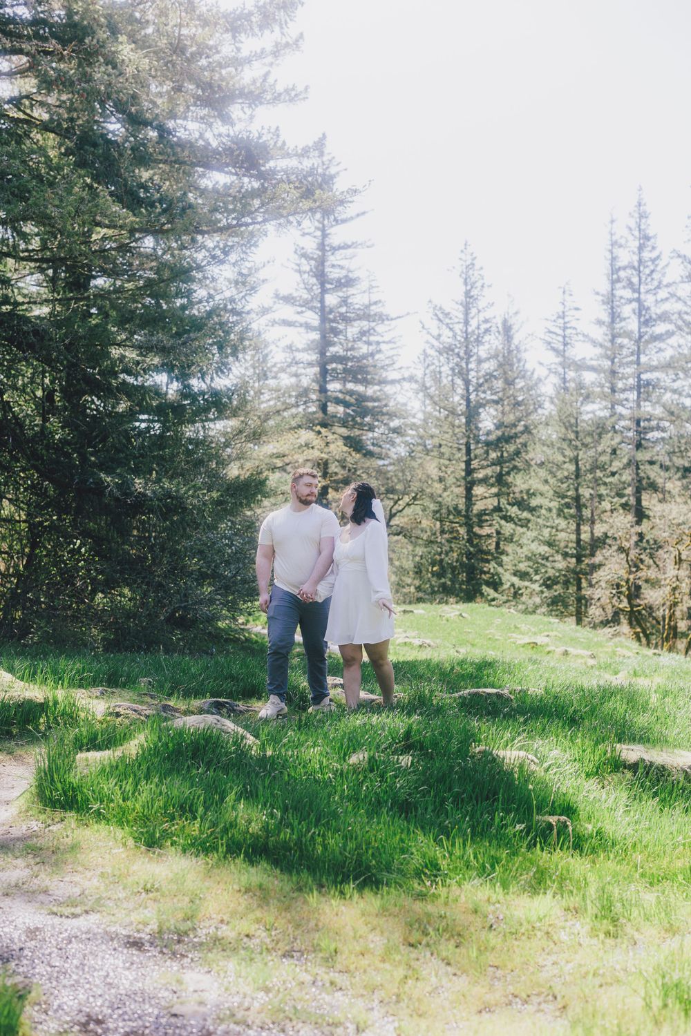 A couple stands together on a hillside path surrounded by evergreen trees.