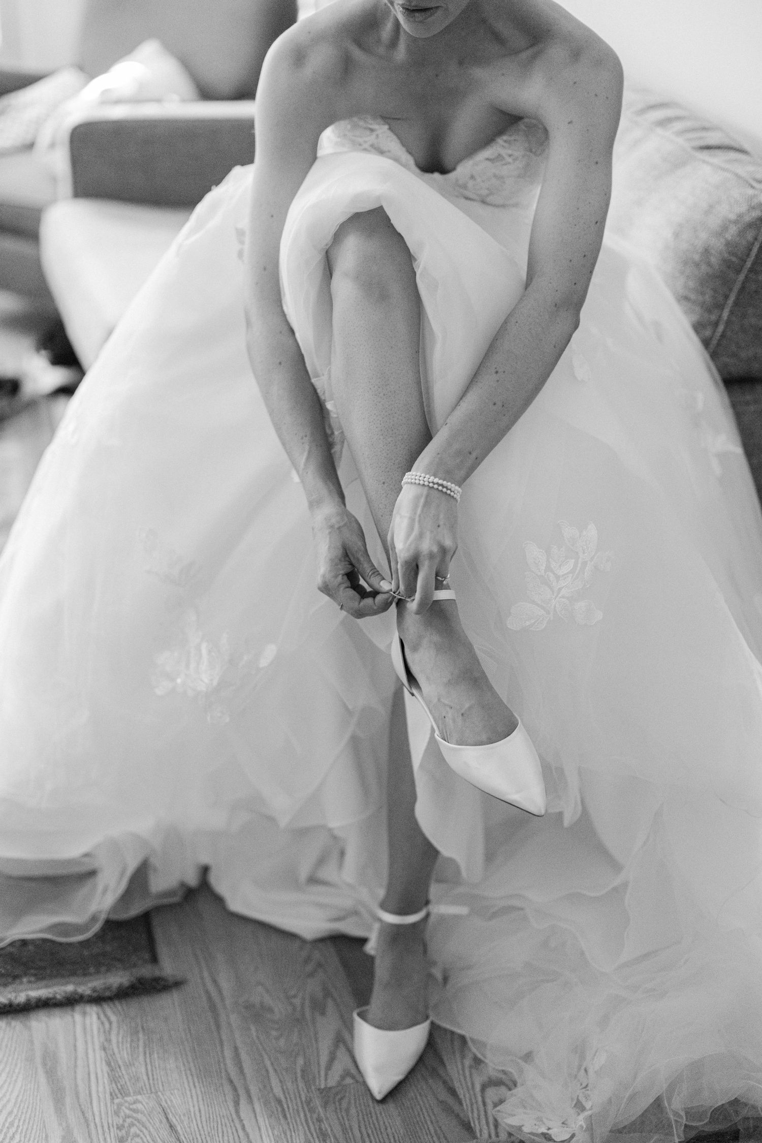 Black and white photo showing someone in a tulle wedding gown fastening delicate white heels while seated.