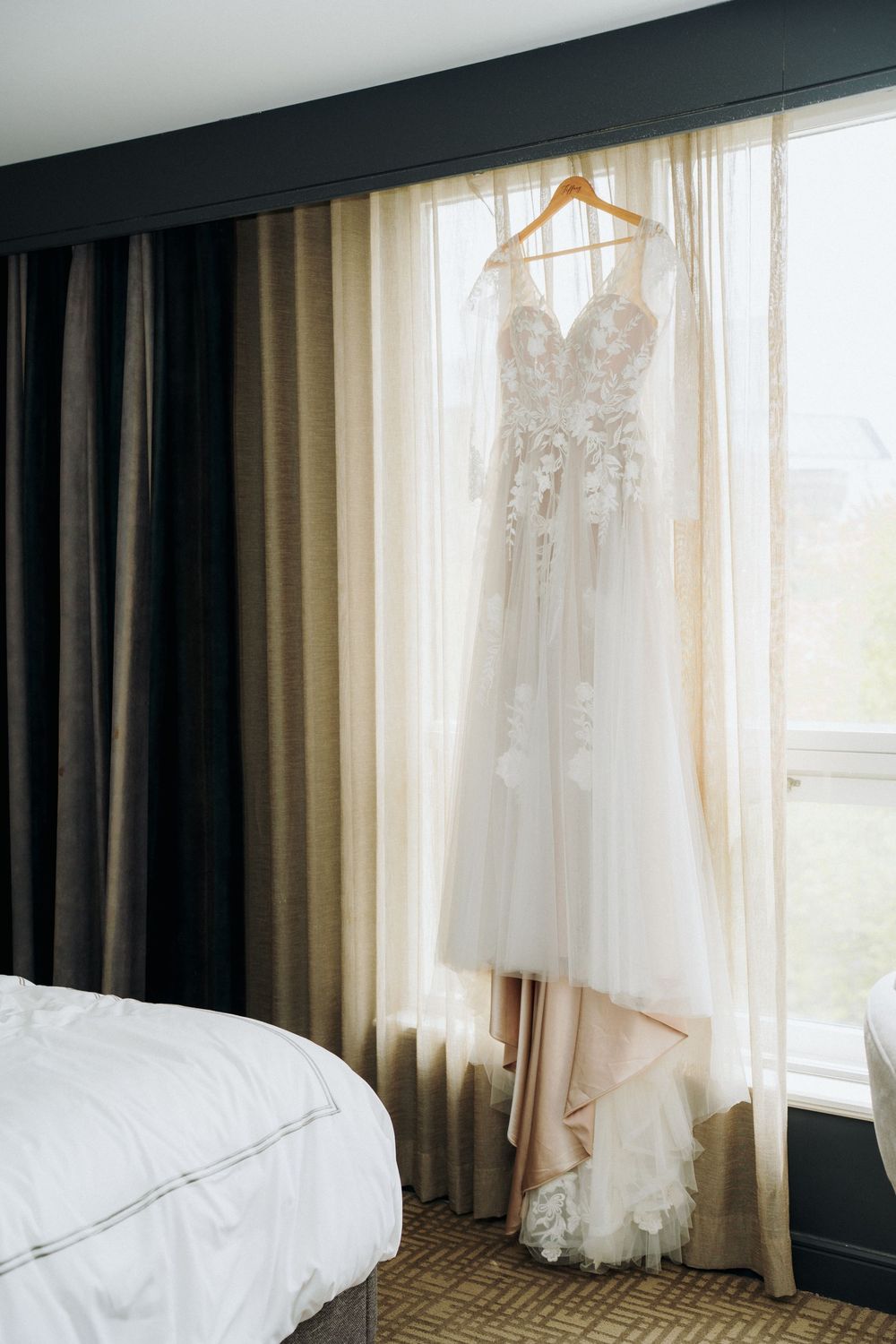 White lace wedding dress hanging by window with sheer curtains and bed visible.