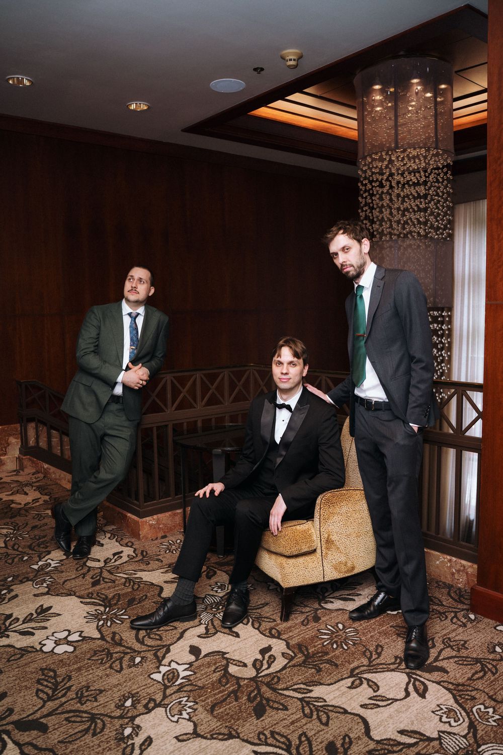 Groomsmen in dark suits examine an ornate floral carpet pattern in a luxury hotel hallway.