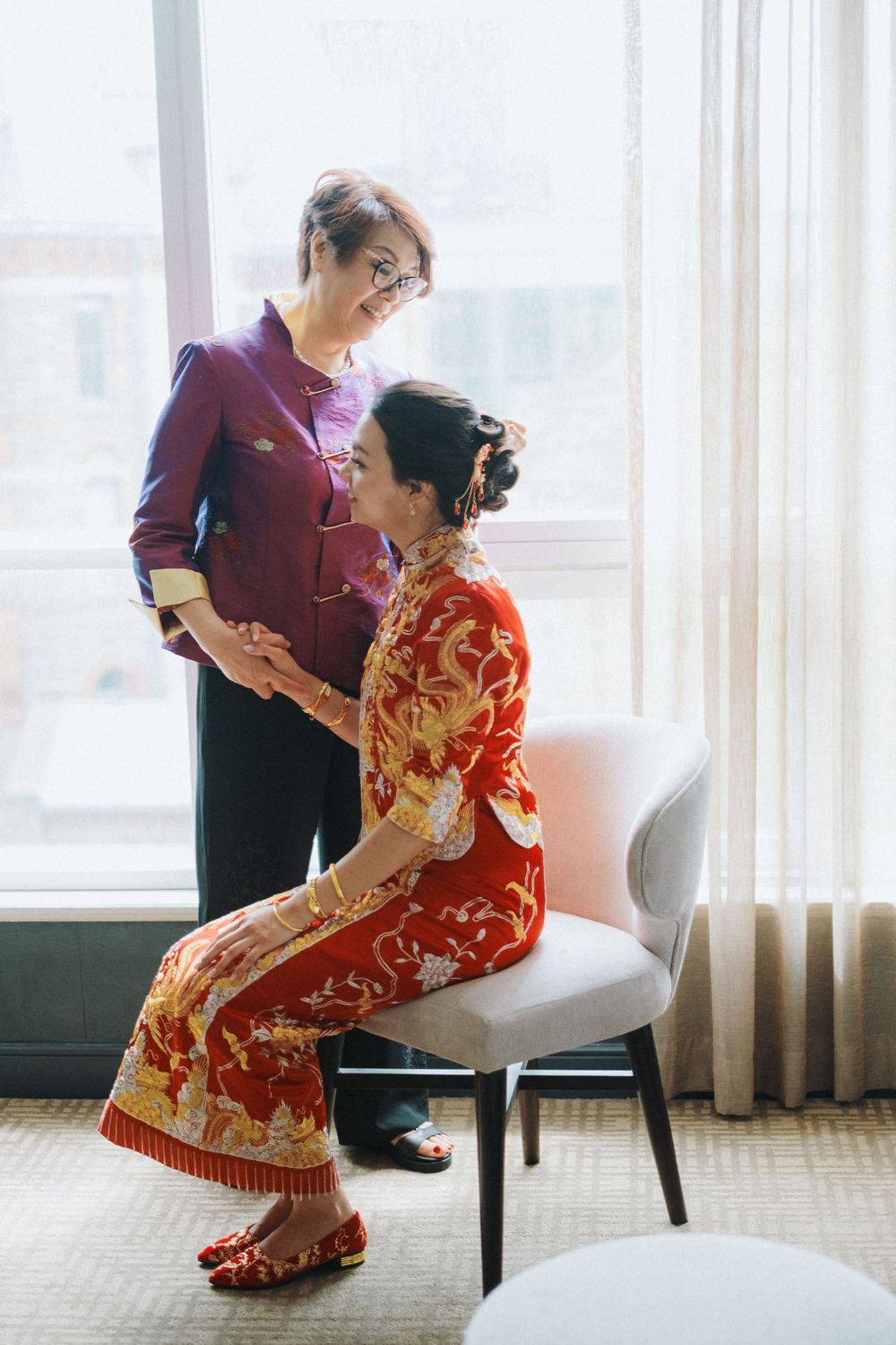 A woman in a traditional red qipao dress is seated elegantly on a chair by a bright window.