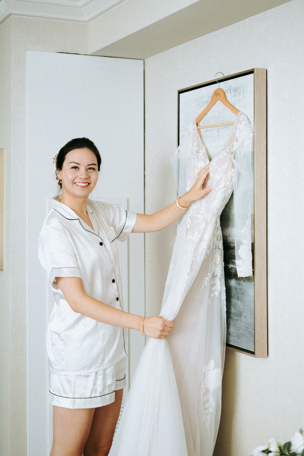 Bride in white pajamas holding wedding dress hanging on display.