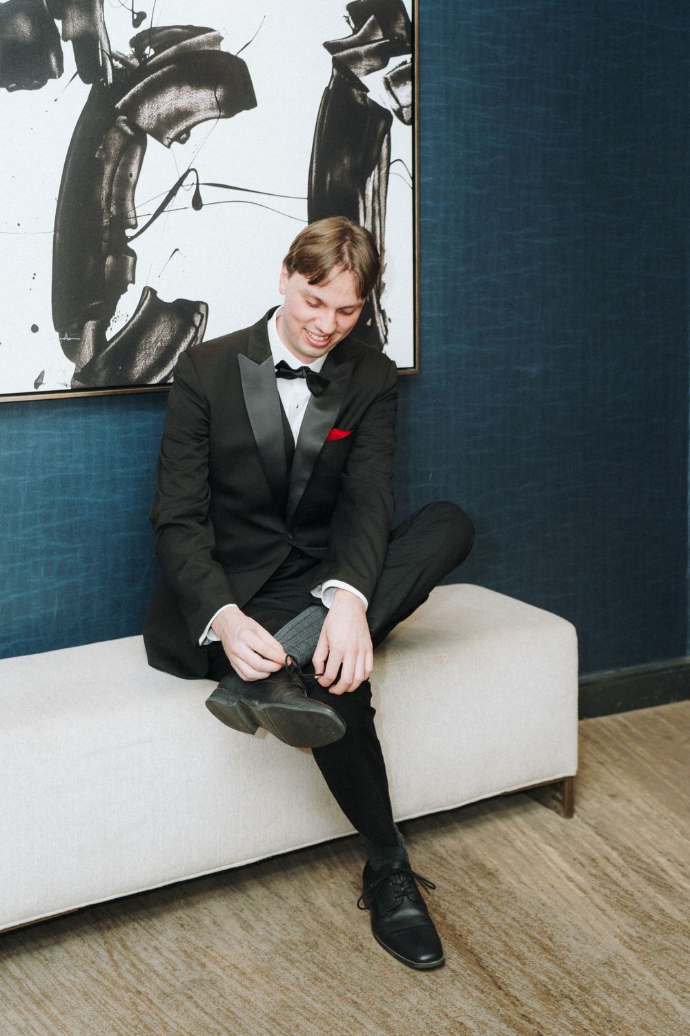 Person in formal black tuxedo sitting on white couch with modern art on wall.