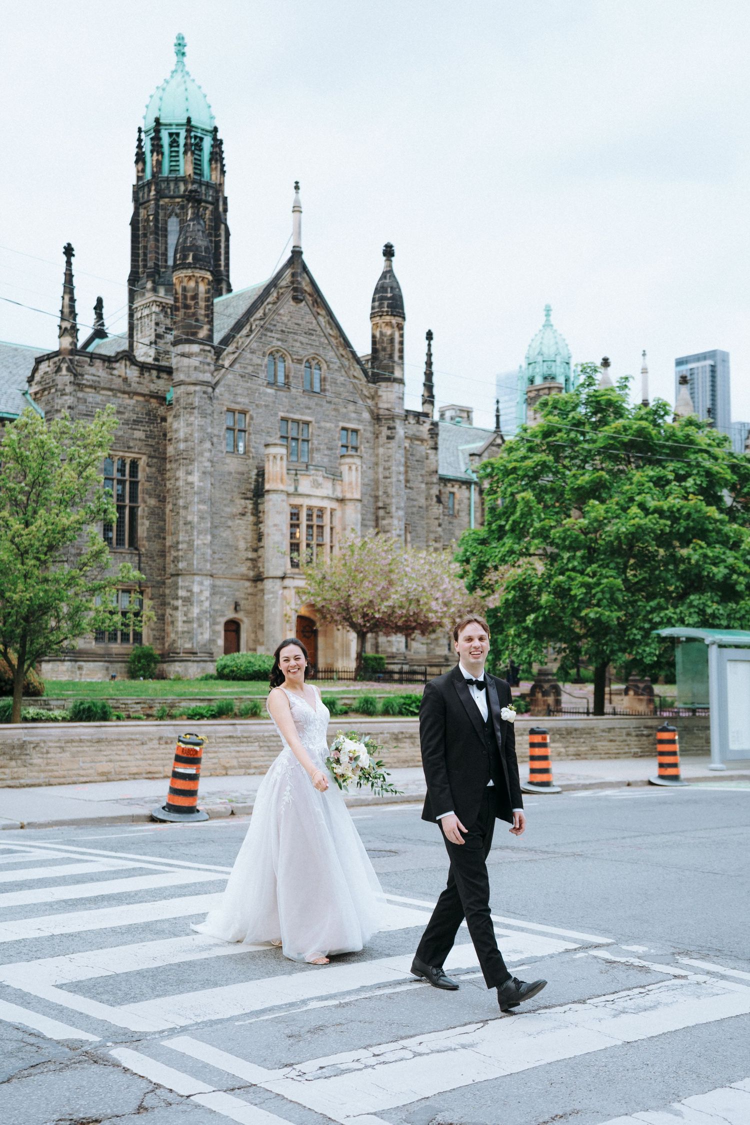 Bride in flowing gown and groom in tuxedo walking past Gothic church architecture.