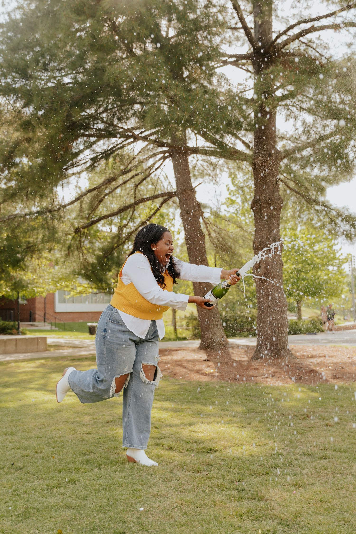A person in ripped jeans and yellow sweater playfully sprays a bottle of champagne in a park with tall pine trees in the background.