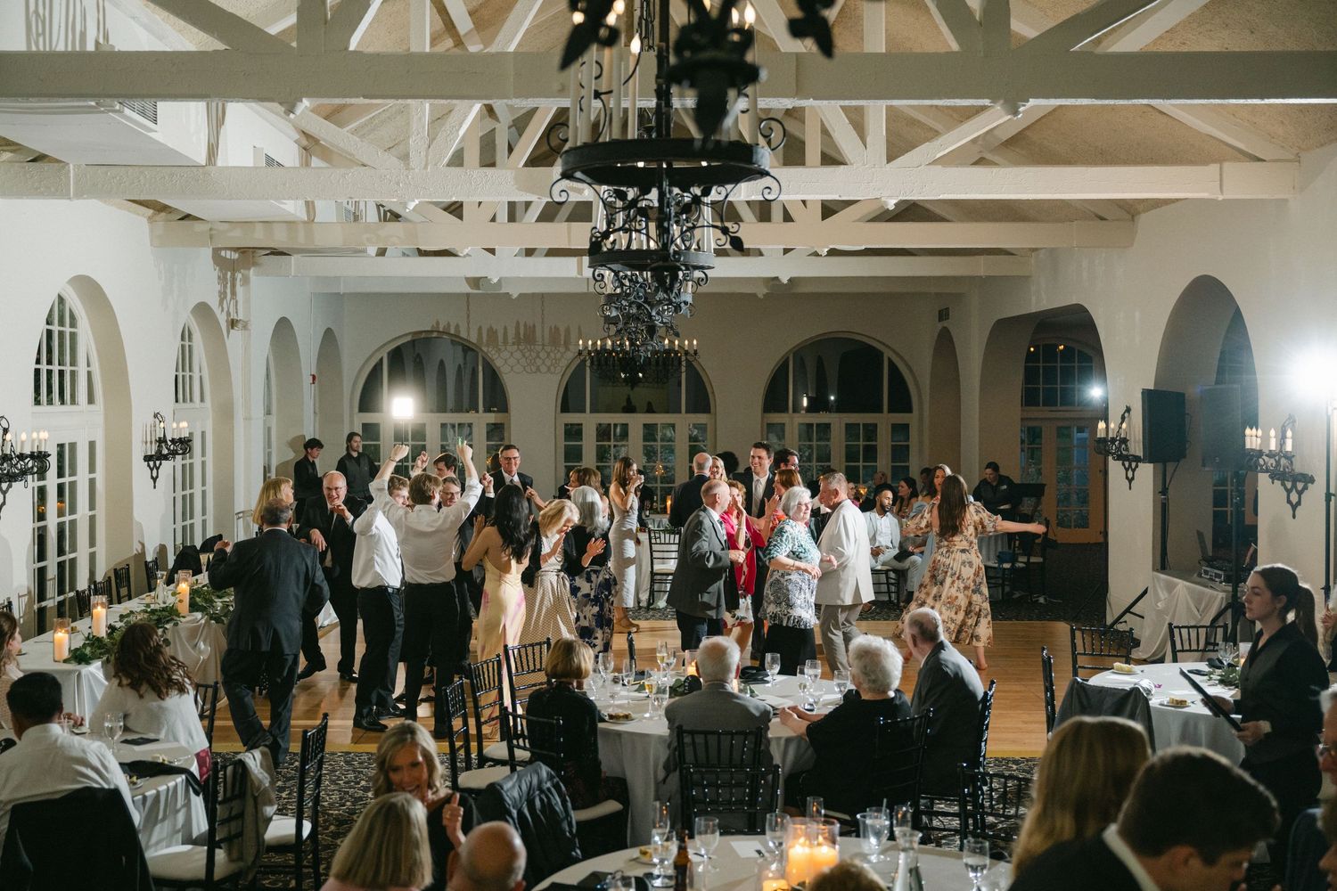 Wedding reception celebration in an elegant white room with exposed beams and people dancing and mingling around tables.