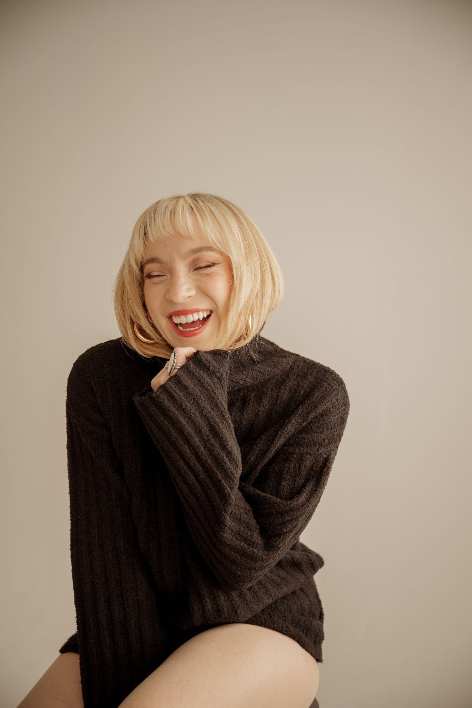 Person wearing a cozy brown sweater laughs joyfully against neutral background.