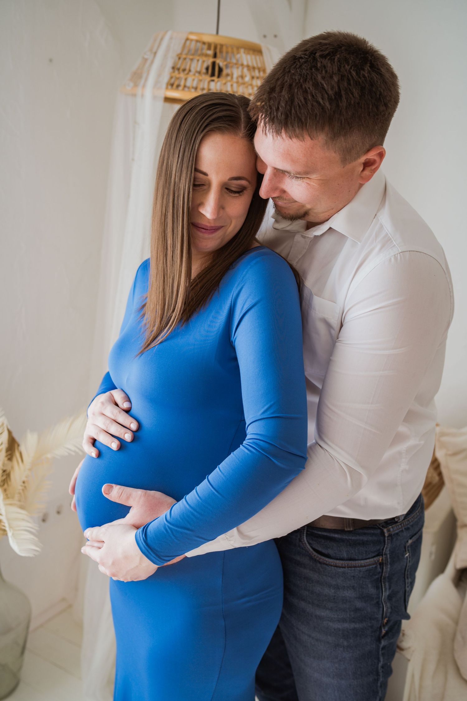A tender maternity photoshoot featuring an expectant couple in a bright room, with the woman wearing a blue fitted dress.