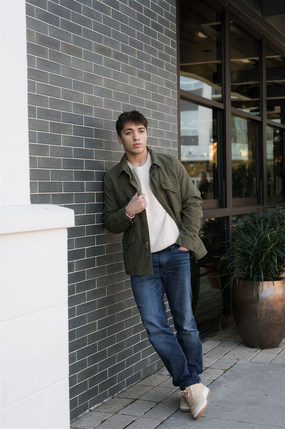 Urban casual fashion look with olive jacket, white t-shirt, blue jeans and sneakers against a gray brick wall exterior.