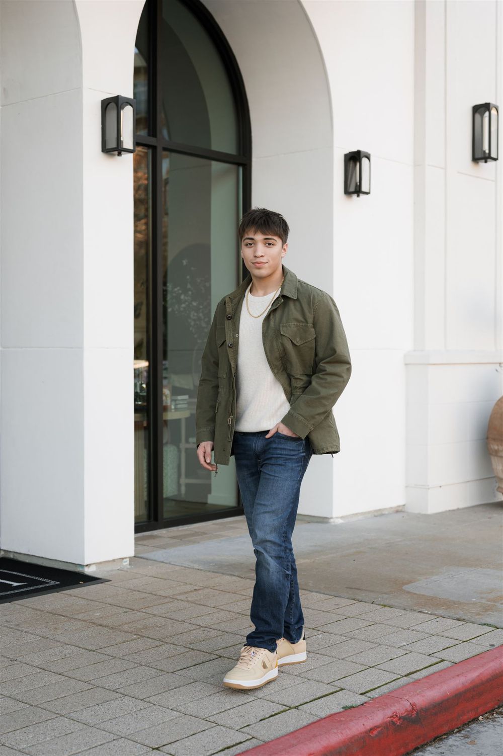 Person in olive military jacket and jeans stands casually in front of white building with arched doorway.