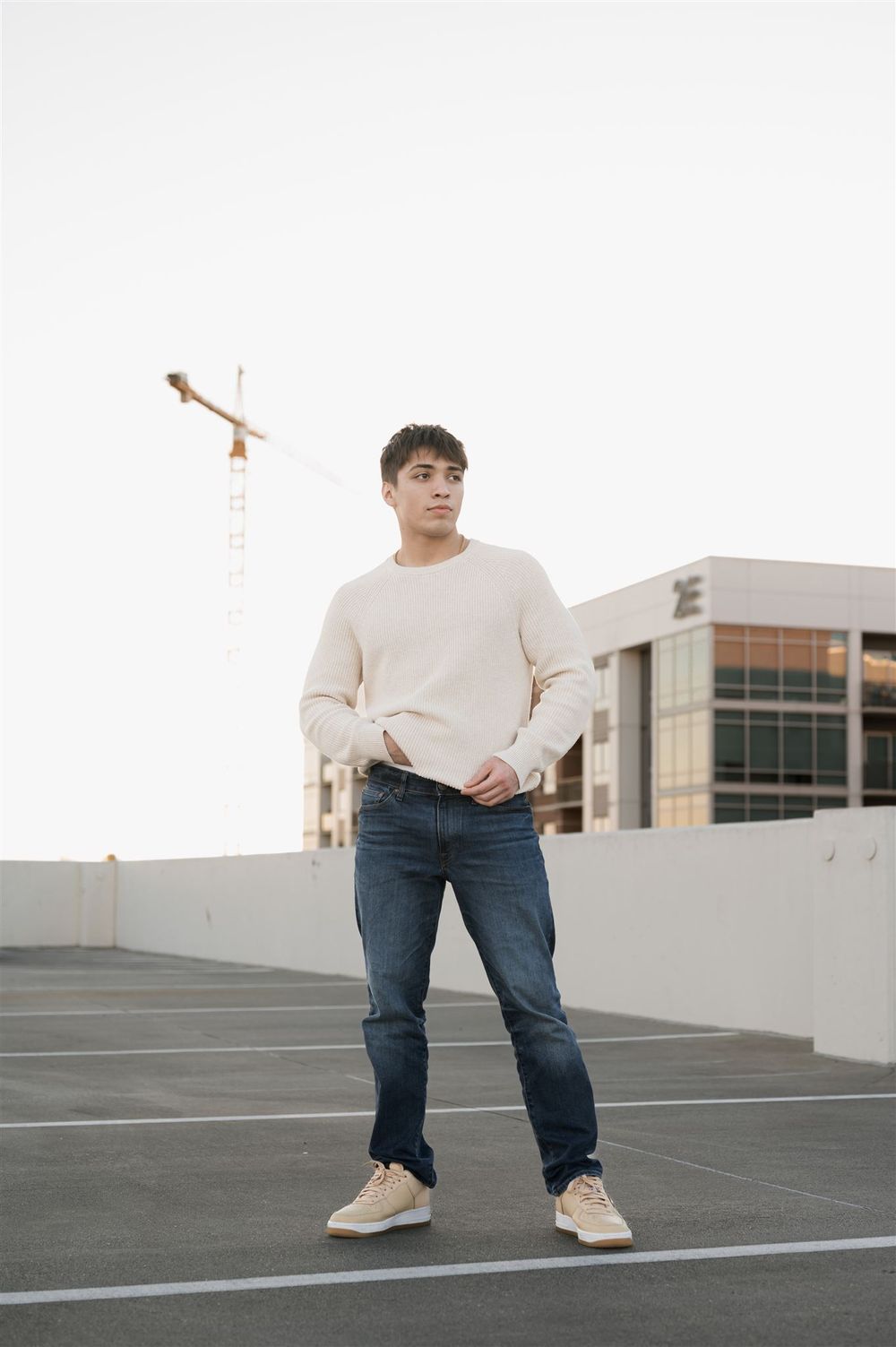 A person standing on a parking garage rooftop wearing a cream sweater, blue jeans and light colored sneakers.
