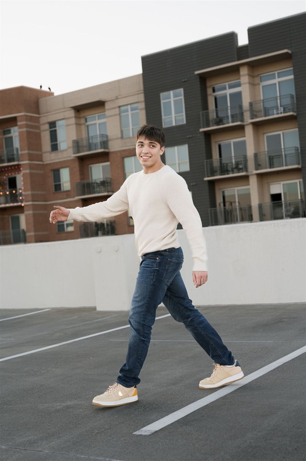 Someone in a cream sweater and blue jeans walking playfully across an empty parking lot with apartment buildings in background.