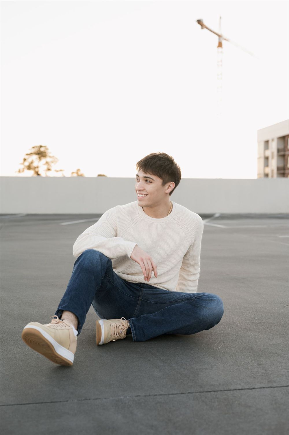 A person wearing a white sweater and jeans sits casually on a rooftop with urban buildings and a crane in the background.