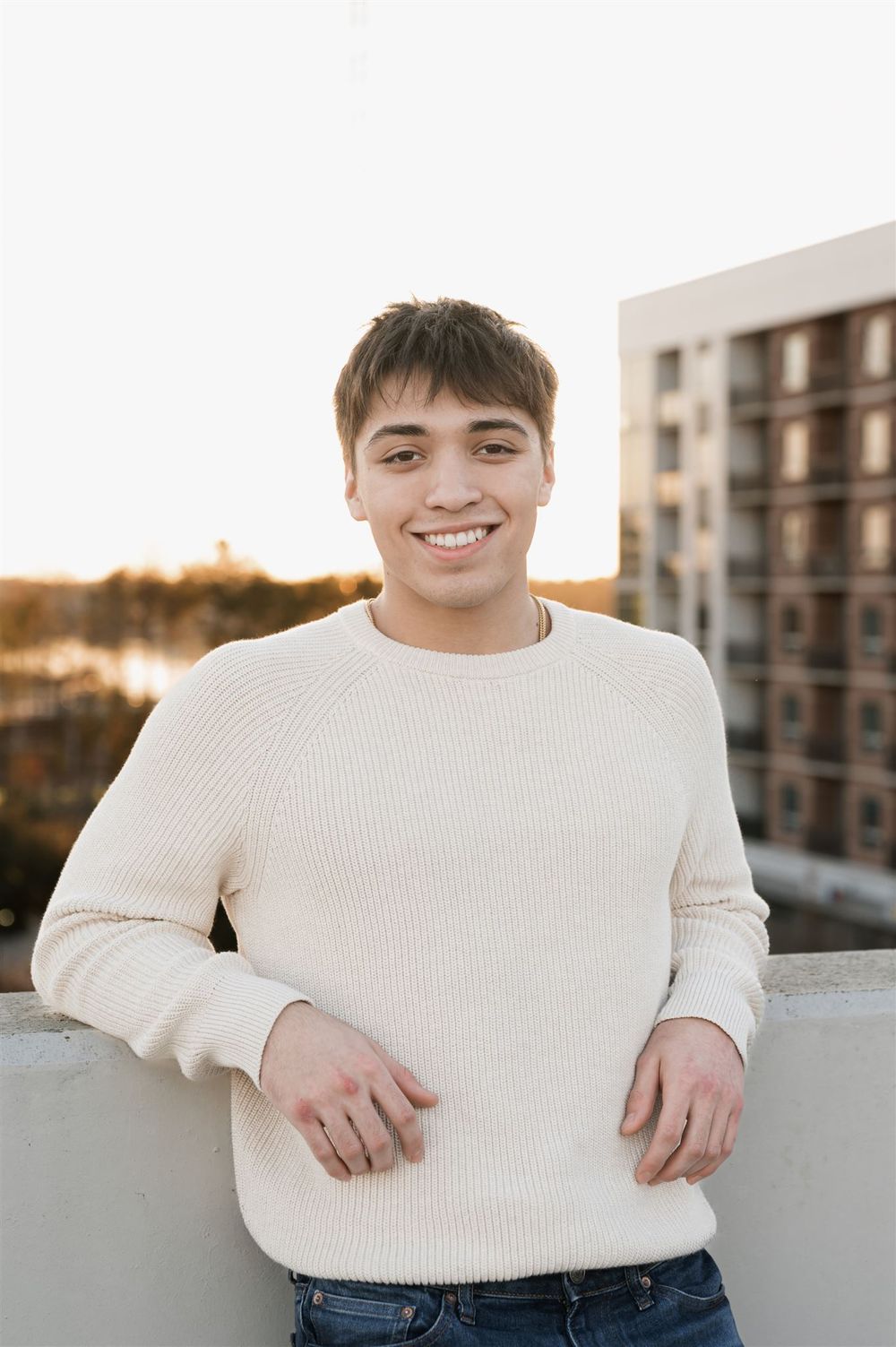 A person wearing a cream ribbed sweater smiles while standing on a rooftop at sunset with an apartment building in background.