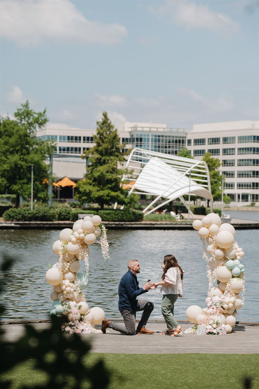 A romantic proposal setup on a dock with balloon arch decorations and office buildings visible across the water.