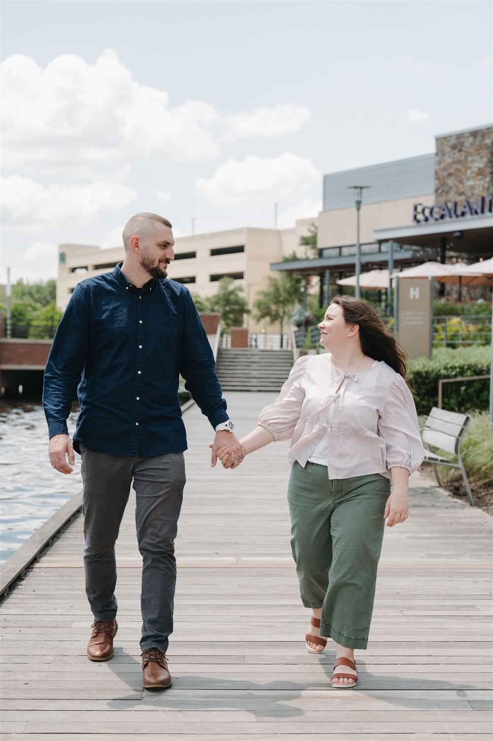 A couple holding hands and walking together on a wooden boardwalk near a shopping center on a sunny day.