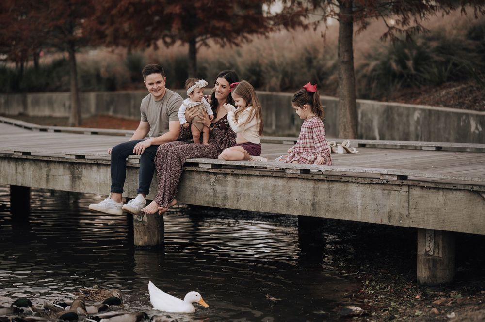 A group sits together on a wooden dock over a pond with a white duck swimming below in the water.