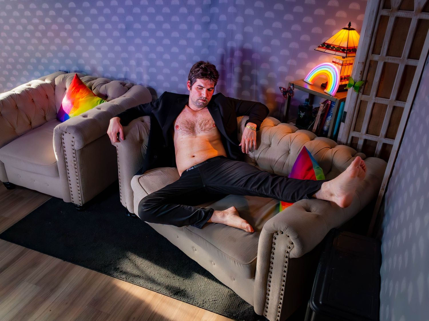Person lounges shirtless on a beige tufted couch in a dimly lit room with ambient lighting and textured walls.