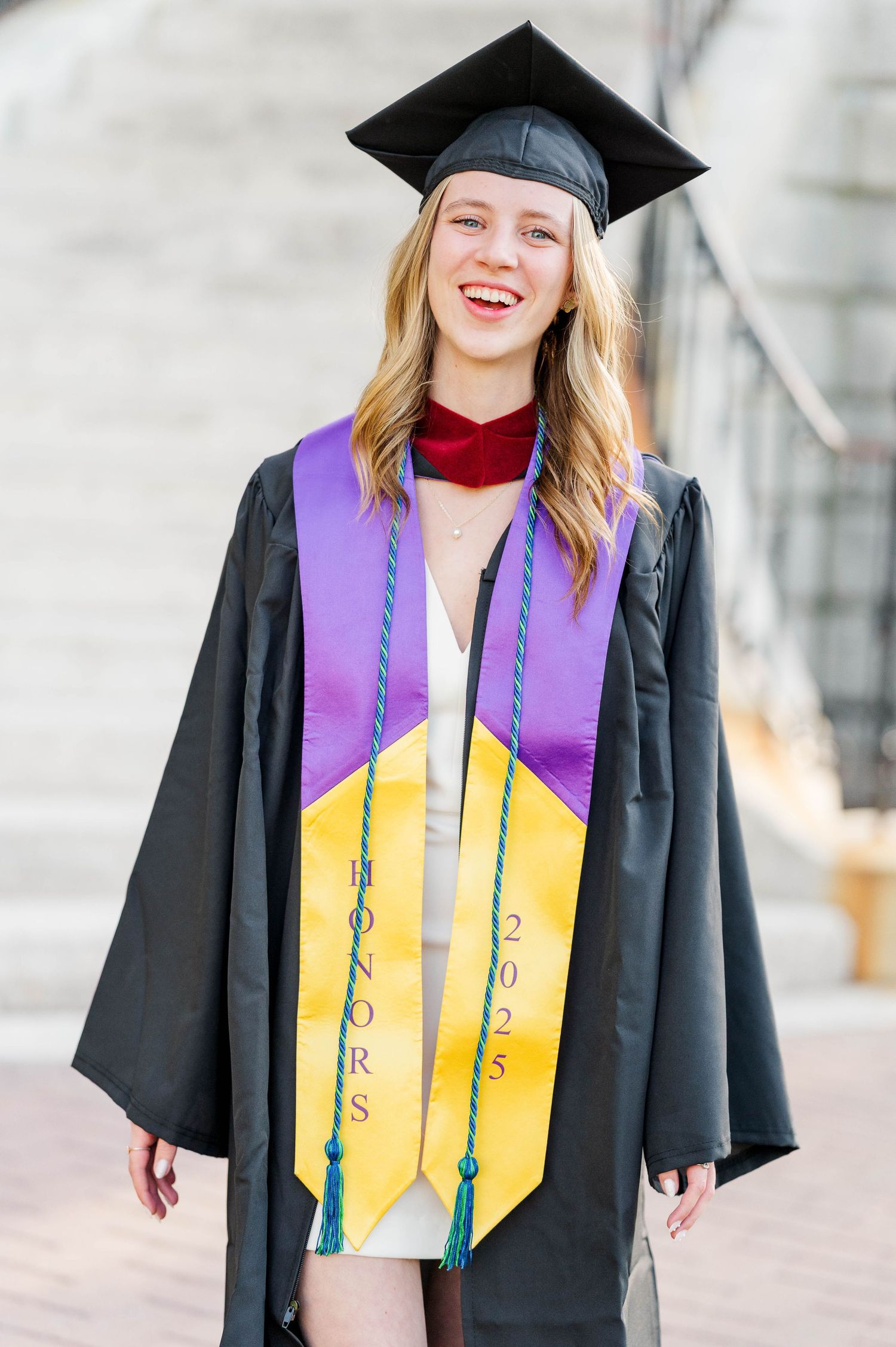 Graduate wearing traditional black cap and gown with purple and gold honors stole celebrating commencement ceremony.