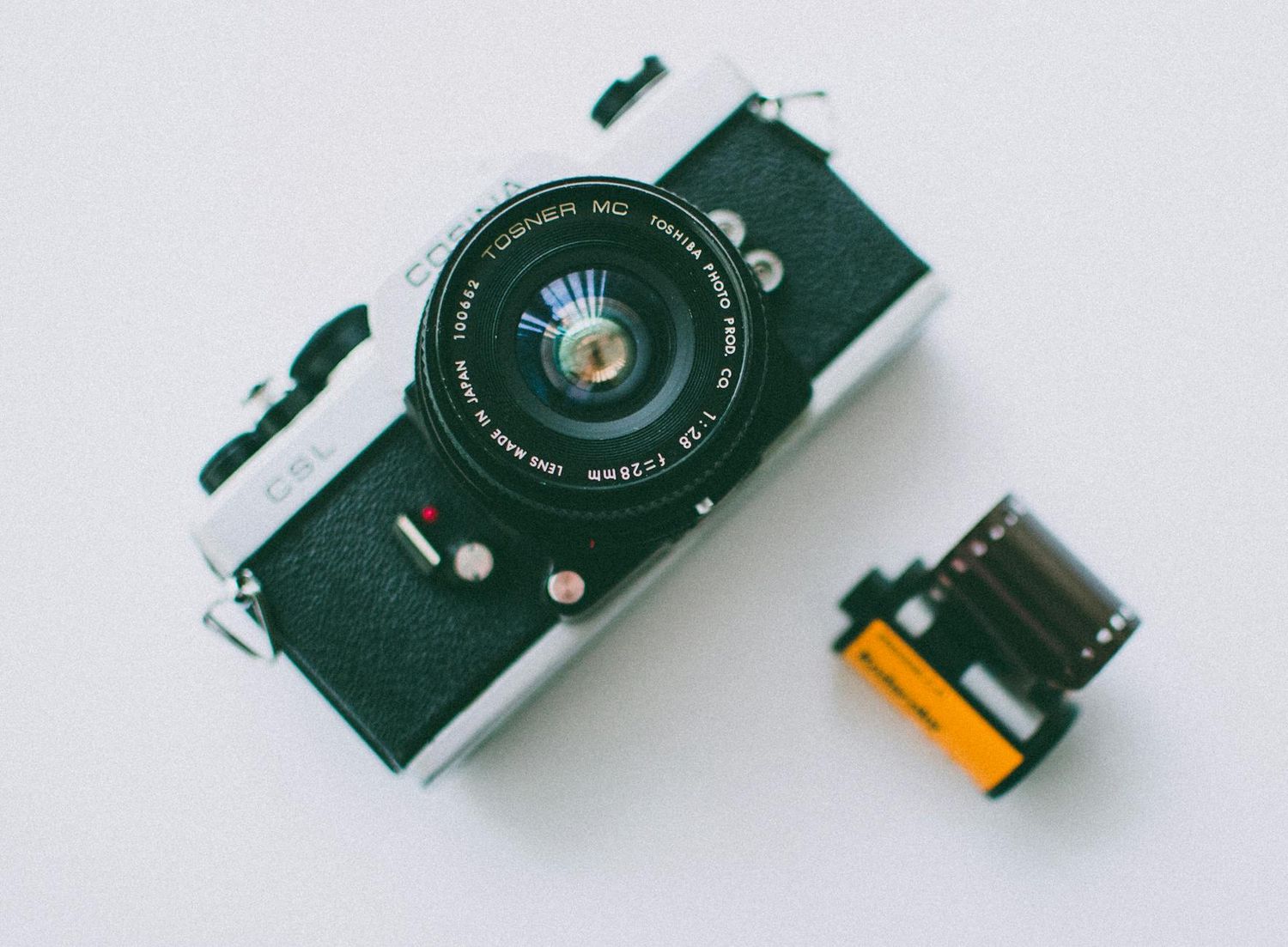 A vintage black film camera sits next to a roll of analog film on a white background.