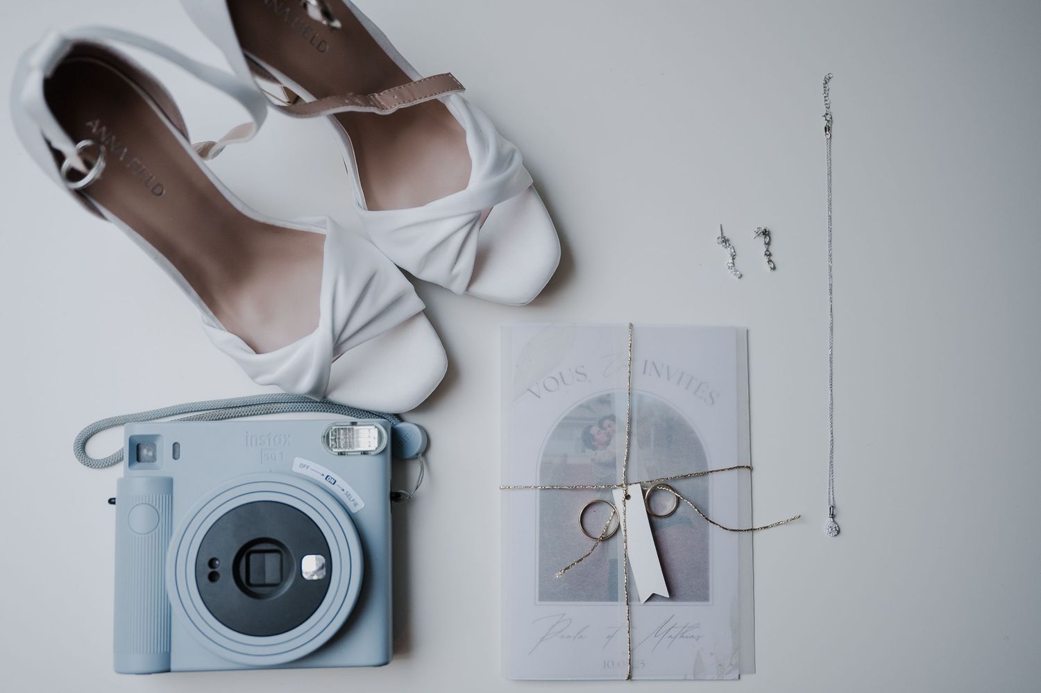 Flat lay photography of white bridal shoes, Instax camera, and wedding rings on a light gray surface.