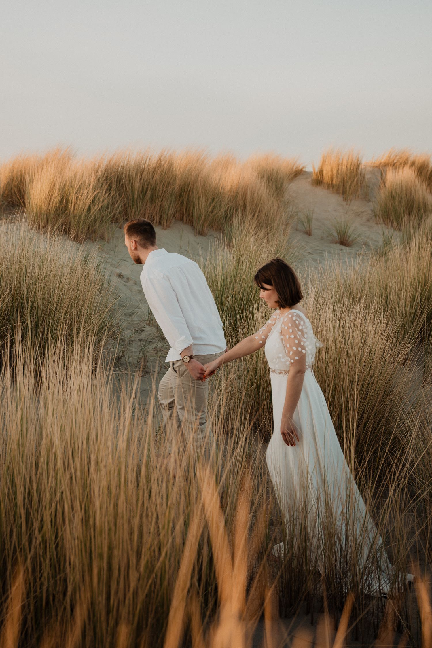 A couple walks through tall golden beach grass at sunset in romantic wedding attire.